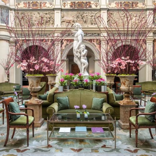 Elegant lounge area in a grand indoor courtyard of the Four Seasons Hotel Firenze, decorated with lush green velvet sofas, wooden chairs, large floral arrangements, and a classical statue in the background.