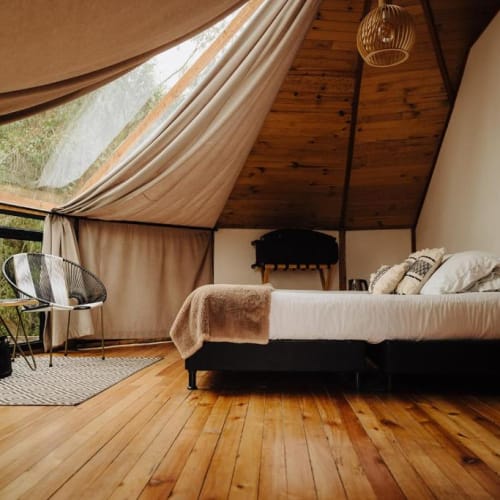 Cozy bedroom in a glamorous camping setup with a bed, side table, two chairs, and large windows with curtains.