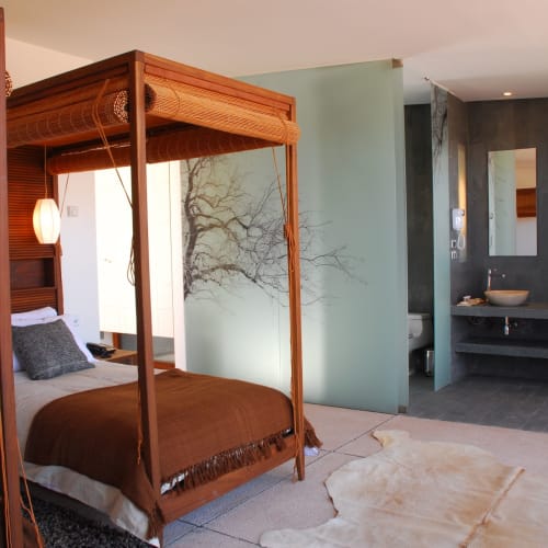 A cozy bedroom with a four-poster bed in a modern, stylish hotel room with a view of a tree through the wall.