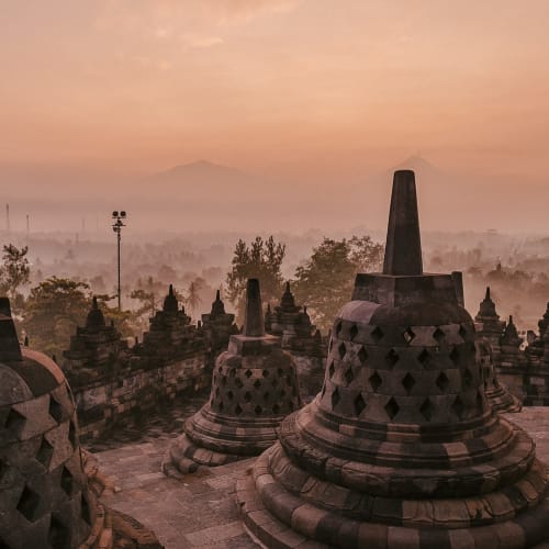 Sunset over Borobudur Temple with multiple stupas in the foreground and mountain in the background, misty atmosphere.