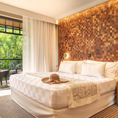 Modernes Schlafzimmer mit großem weißem Bett, strukturiertem Holz-Akzentwand, kleinem Holztisch und Balkon mit Blick auf üppige grüne Palmen.