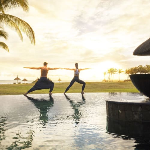 Ein Mann und eine Frau üben Yogaposen neben einem Infinity-Pool bei Sonnenuntergang mit Palmen und einer Grasfläche im Hintergrund.