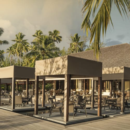 Außenbereich eines Restaurants in einem Resort mit überdachten Sitzbereichen, umgeben von Palmen und einem großen Gebäude im Hintergrund unter bewölktem Himmel.
