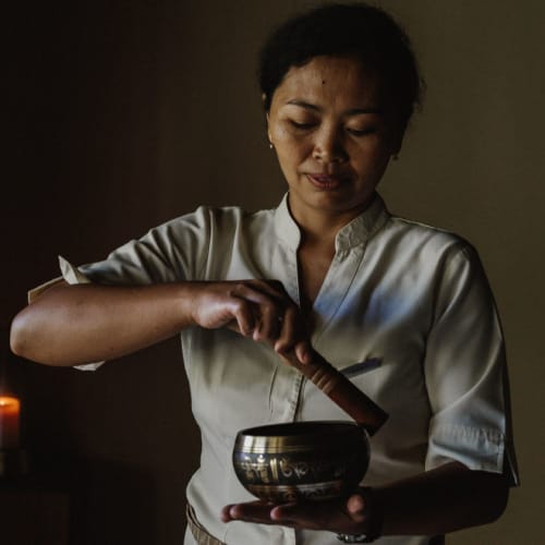 A woman performs a traditional sound healing ritual using a singing bowl in a dimly lit space.
