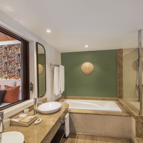 Modern bathroom with a green accent wall, a bathtub, a glass-enclosed shower, dual sinks, and a window opening into a bedroom with patterned wallpaper and orange cushions.