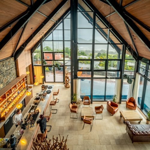 Innenraum einer geräumigen Lodge mit hoher Holzdecke, großen Glasfenstern mit Blick auf einen Pool und Außenbereich, mit Barbereich und mehreren Sitzgelegenheiten einschließlich Stühlen und Sofa.