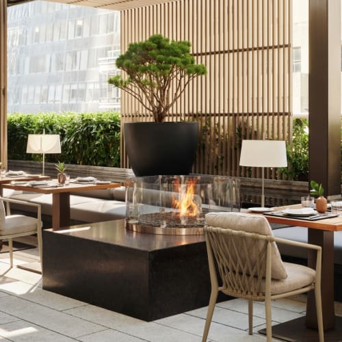 A stylish outdoor restaurant with a fire feature in the center, surrounded by dining tables, chairs, and potted plants.
