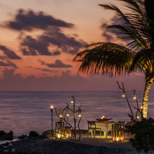Romantisches Dinner am Strand bei Sonnenuntergang mit Tisch, Stühlen und Dekorationslichtern unter einer Palme in Maalifushi, Malediven.