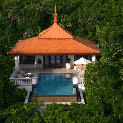 A tropical villa with a traditional orange pagoda-style roof surrounded by dense green foliage, featuring a private rectangular swimming pool and a patio with lounge chairs and an umbrella.