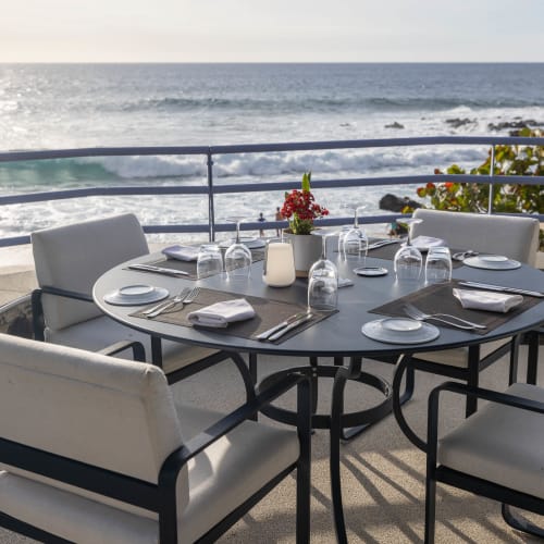 Esstisch im Freien mit vier weißen gepolsterten Stühlen mit Blick auf den Ozean und die felsige Küste bei klarem Himmel.