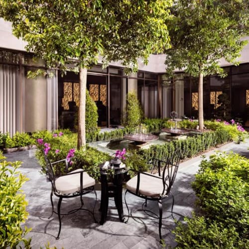 A peaceful hotel garden with two chairs and a small table, surrounded by lush green bushes, trees, and colorful flowers, with modern glass building windows in the background.