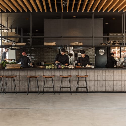 Moderne Restaurantküche mit Holzdecke, fünf Barhockern und fünf Köchen, die an einem langen, hellen Fliesentisch verschiedene Aufgaben erledigen.