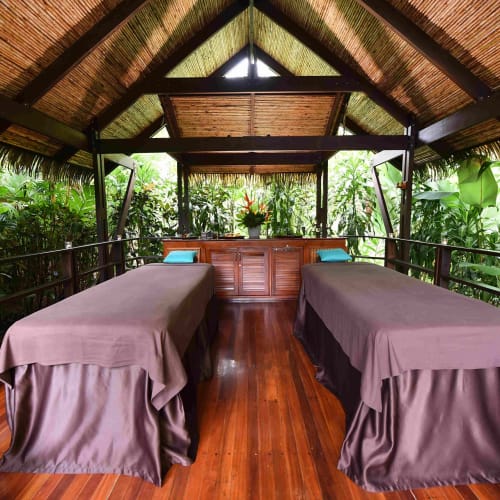 A wellness area in a tropical outdoor spa with two massage beds, surrounded by lush greenery and built with natural wood and thatched roof.