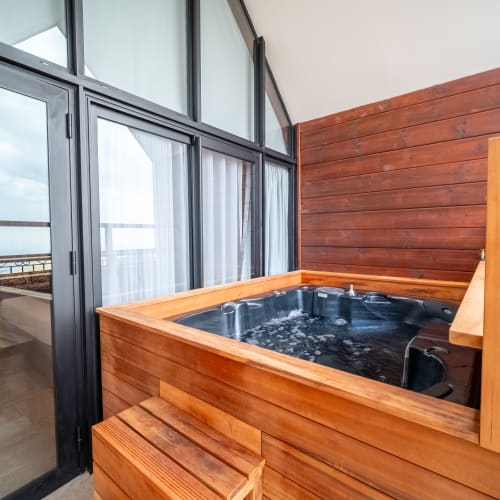 Ein hölzerner Whirlpool mit sprudelndem Wasser in einem Raum mit großen Glastüren und einem Balkon mit Blick auf Strand und Meer.