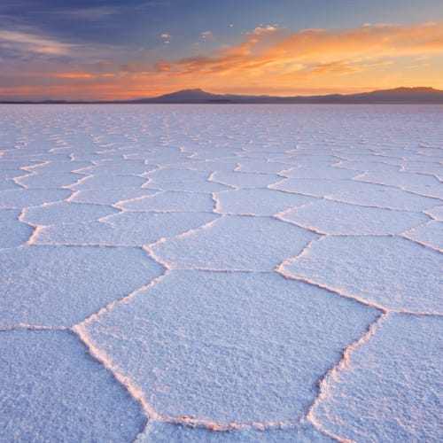 Expansive salt flats at sunrise with hexagonal salt crusts and distant mountains under a colorful sky.