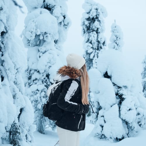 Eine Person mit weißer Mütze, schwarzer Jacke mit Fellkapuze und weißer Hose steht zwischen schneebedeckten Bäumen in einer Winterlandschaft.