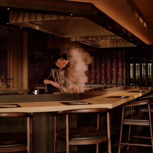 A chef cooking at the Aman Nai Lert Bangkok Aman Hiori restaurant, with steam rising from the dish, in a modern, warmly lit interior.