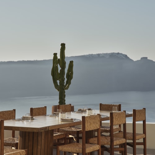 Outdoor dining table with wooden chairs and place settings, overlooking a calm sea with distant hills and a cactus plant in the background.