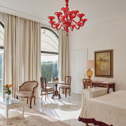 Elegantes Hotelzimmer mit großen Fenstern, rotem Kristallleuchter, antiker Einrichtung und Blick auf einen Garten und Sitzbereich im Freien.