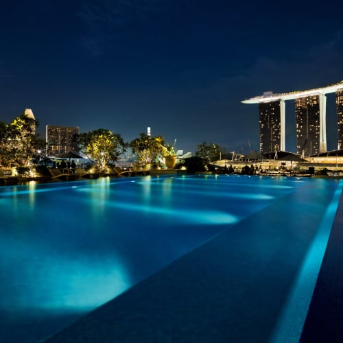 Ein nächtlicher Blick auf ein beleuchtetes Schwimmbecken mit der Skyline Singapurs, einschließlich des ikonischen Marina Bay Sands Hotels, im Hintergrund.