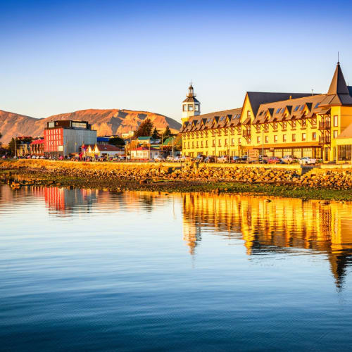 Ein malerischer Sonnenuntergangsblick auf Puerto Natales mit bunten Gebäuden, die im ruhigen Wasser reflektiert werden, und entfernten Bergen.