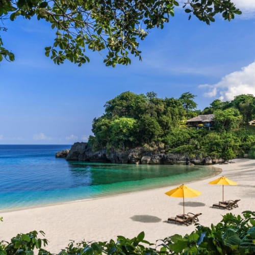 A tropical beach with white sand, turquoise water, and yellow umbrellas with lounge chairs, surrounded by lush green trees and a clear blue sky.