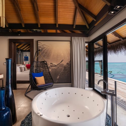 Luxuriöses Badezimmer in einer Wasser-Pool-Villa mit großem runden Whirlpool, Hängesessel mit Kissen und Panoramafenstern mit Blick auf den Ozean und die private Terrasse in einem tropischen Resort.