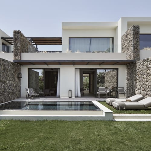 Moderne Villa mit Steinmauern und privatem rechteckigem Pool, zwei Sonnenliegen und Sitzstühlen auf einer überdachten Terrasse vor einem Rasen.