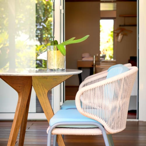 Blaue gepolsterte Stühle mit Rückenlehnen aus geflochtenem Seil um einen Holztisch auf einer Holzterrasse, darauf eine Glasvase mit rosa Orchidee. Eine offene Tür führt zu einem warm beleuchteten Innenraum mit Holzbalken.