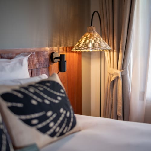Gemütliche Schlafzimmerecke mit einem Bett, dekorativen Kissen und einer Rattan-Wandlampe neben einem verhängten Fenster.