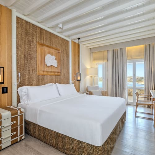 Bright bedroom with a large bed, wooden accents, and a view of the sea outside the windows.