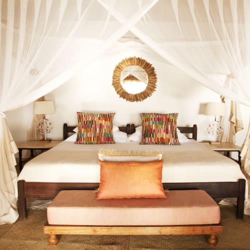 Luxuriöses Schlafzimmer mit großem Bett, bunten Kissen und einem Baldachin aus weich fließenden weißen Vorhängen in einer tropischen Umgebung.
