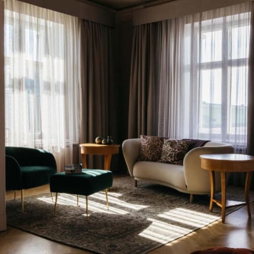 Cozy living room with beige sofa, dark green armchair and ottoman, wooden side tables, large windows with sheer and dark curtains, and sunlight casting shadows on a patterned rug.