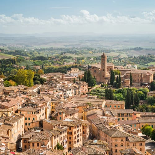 A panoramic view of Siena, Italy, showcasing the historic medieval buildings with terracotta roofs and a distant church tower amidst green rolling hills under a partly cloudy sky.