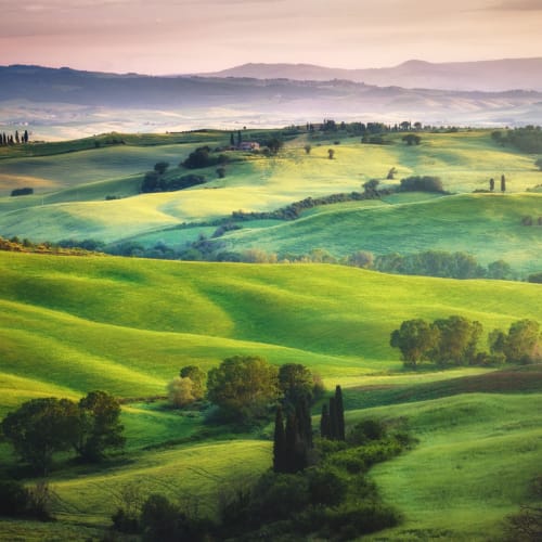 Rolling green hills with scattered trees and distant mountains in Tuscany, Italy, during daytime.