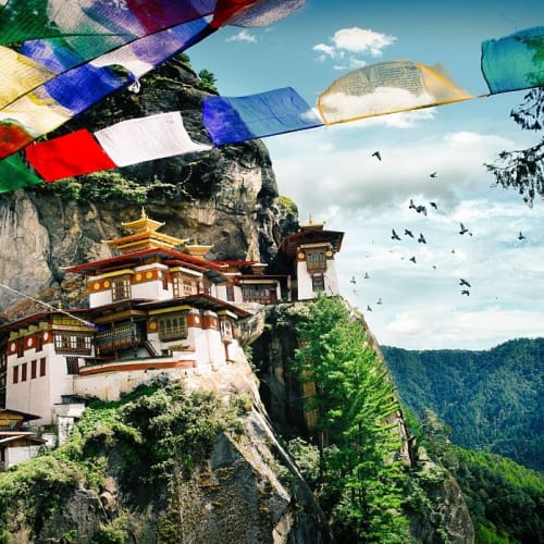 Tiger's Nest Kloster auf einer steilen Klippe in Bhutan, mit Gebetsfahnen im Vordergrund und grünen Bergen im Hintergrund.