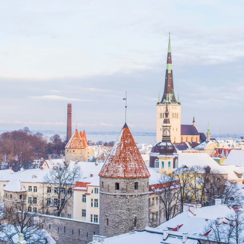 Schneebedeckte Dächer und mittelalterliche Türme in Tallinn, Estland, mit dem markanten Turm der St.-Olavs-Kirche im Hintergrund unter bewölktem Himmel.