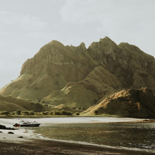 Ein großer Berg mit grünen Hängen erhebt sich hinter einem ruhigen Fluss oder See, mit einem kleinen Boot am dunklen, felsigen Ufer.