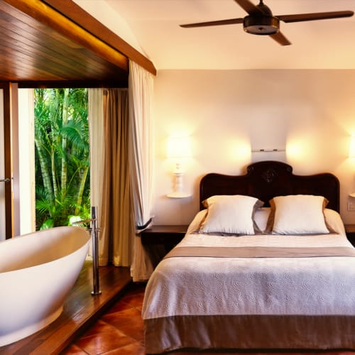Ein gemütliches Hotelzimmer mit einem Doppelbett, zwei Kissen, Nachttischlampen, Deckenventilator und einer ovalen Badewanne neben einem Fenster mit Blick auf grüne tropische Pflanzen.