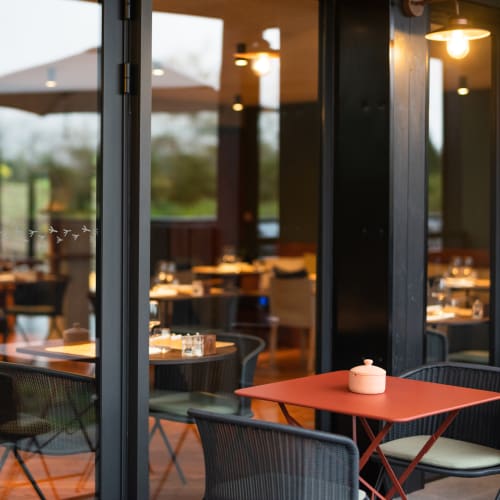 Leere Außen Tische und Stühle eines Restaurants mit Glastüren, die den sanft beleuchteten Innenbereich mit Kerzen und warmem Licht spiegeln.