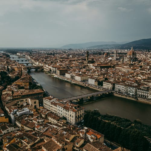 Luftaufnahme von Florenz, Italien, mit dem Arno-Fluss und mehreren Brücken sowie den historischen Gebäuden der Stadt, darunter die markante Kuppel der Kathedrale von Florenz, unter bewölktem Himmel.