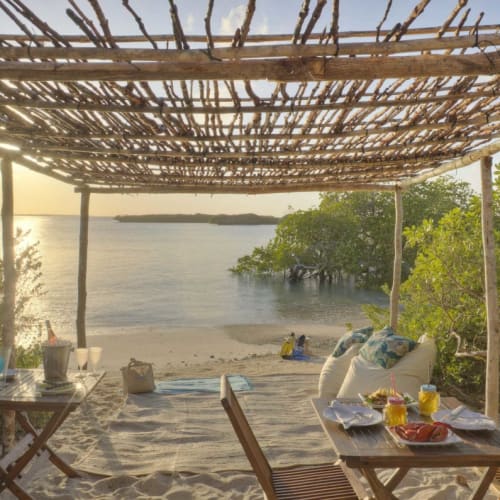 Gemütlicher Außenbereich auf einem sandigen Strand unter einer rustikalen Holzkonstruktion, mit Blick auf den Ozean und grüne Büsche.