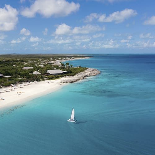 Ein Segelboot fährt nahe einem weißen Sandstrand mit üppigem Grünland und über Wasser liegenden Bungalows im Hintergrund.