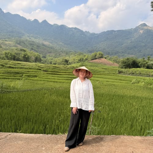 A woman wearing a traditional conical hat stands on a path in front of lush green rice terraces with forested mountains and a partly cloudy sky in the background.