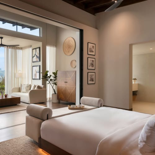 Modernes Schlafzimmer mit Blick auf die Natur durch große Glastüren, die zu einem Sitzbereich mit Sofa, Couchtisch und dekorativer Wandkunst führen.