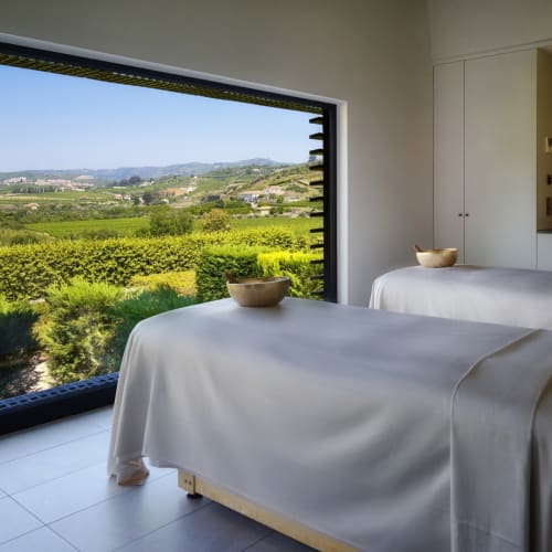 A tranquil spa treatment room with two massage tables covered in white sheets, facing a large window overlooking lush green vineyards and rolling hills in Portugal.