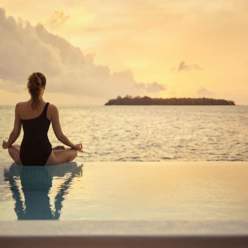 A woman practicing meditation by the infinity pool with the ocean and sunset in the background.