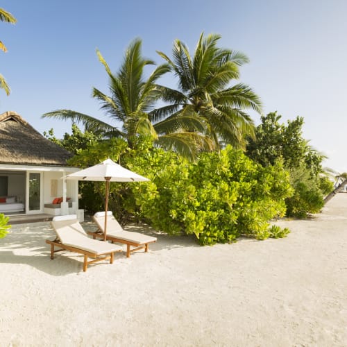 Zwei hölzerne Liegestühle unter einem weißen Sonnenschirm am sandigen Strand neben einer tropischen Villa, umgeben von Palmen und grünen Büschen unter klarem blauem Himmel.