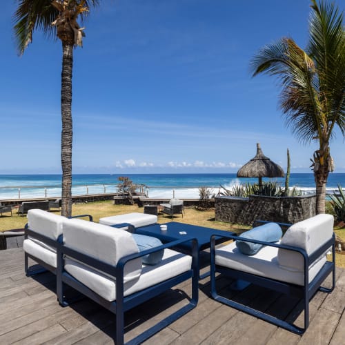 Draußen sitzbereich mit gepolsterten Stühlen und einem Glastisch auf einem Holzdeck mit Blick auf den Ozean und einen Sandstrand mit Palmen unter klarem blauem Himmel.