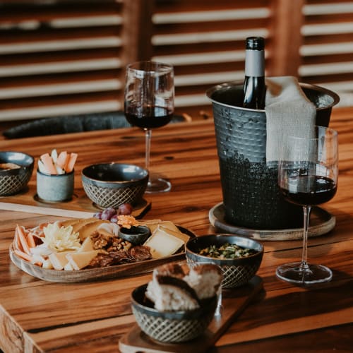 Ein Holztisch mit einem Weinkühler mit einer Flasche, zwei Gläsern Rotwein, einer Käse- und Snackplatte sowie mehreren kleinen Schalen mit Brot und anderen Beilagen.